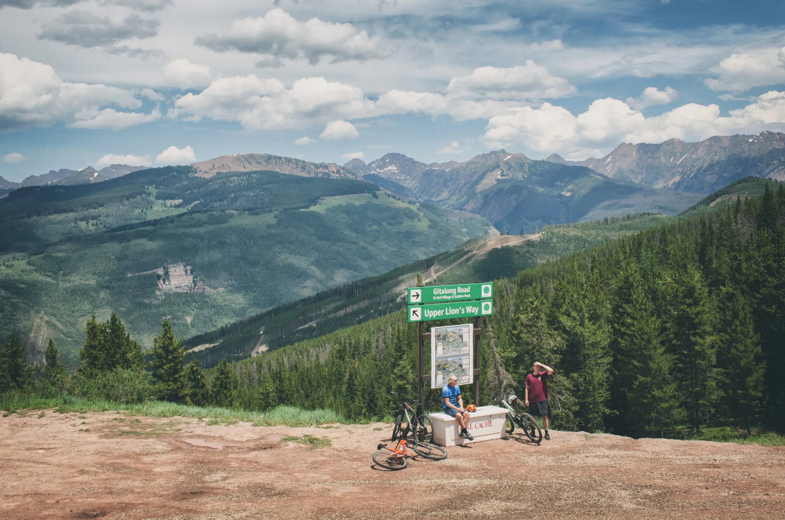 Two mountain bikers rest by trail signs on a ridge overlooking the Colorado high country