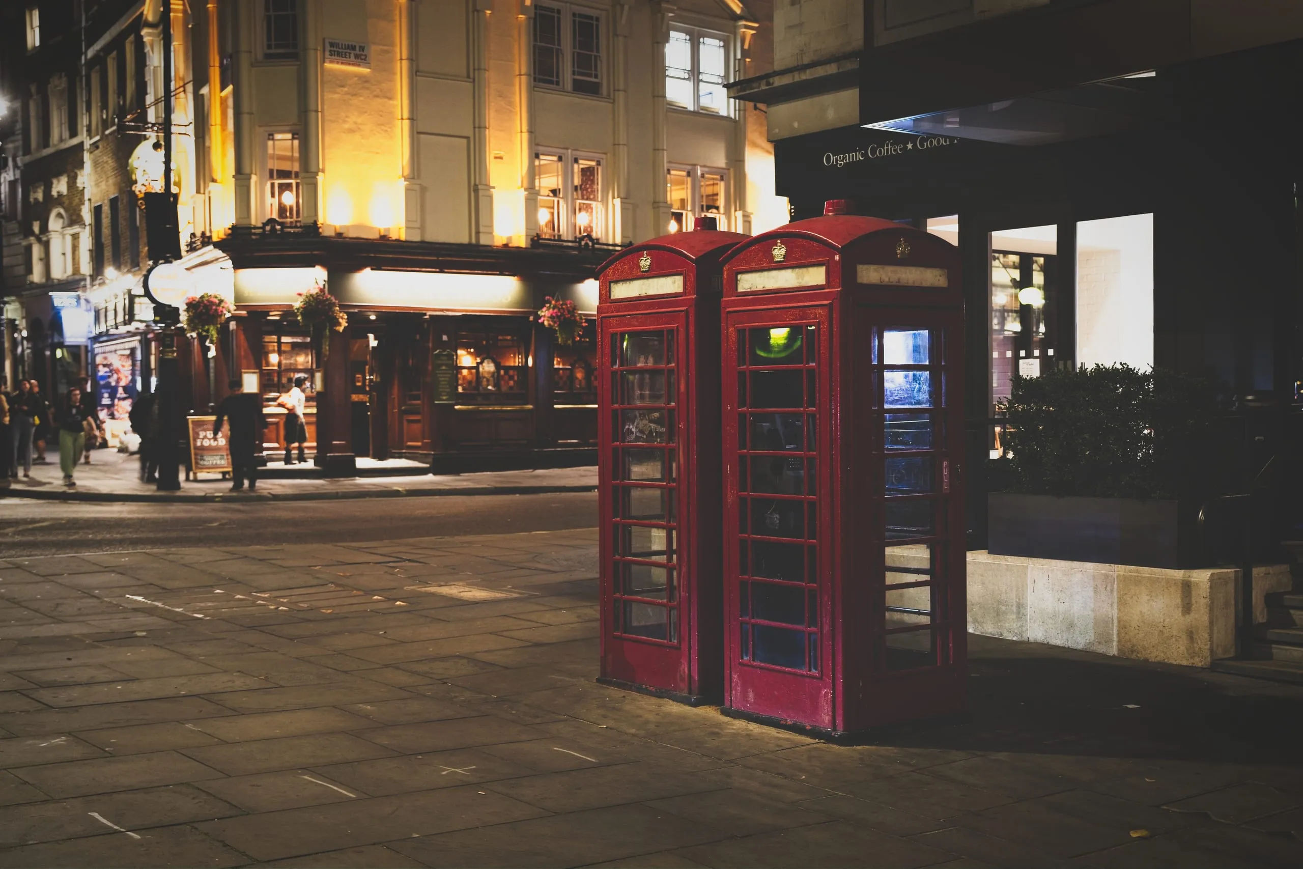 A night photograph of two classic red telephone boxes on a London street corner near William IV Street, with a warmly lit pub in the background.